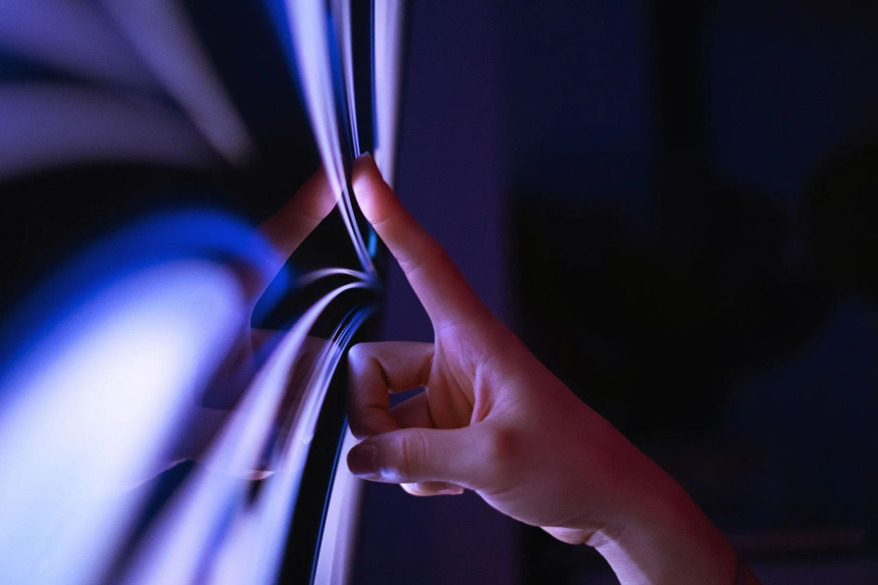 An index finger reaching out to touch a glass surface with streaks of purple light and a dark background.