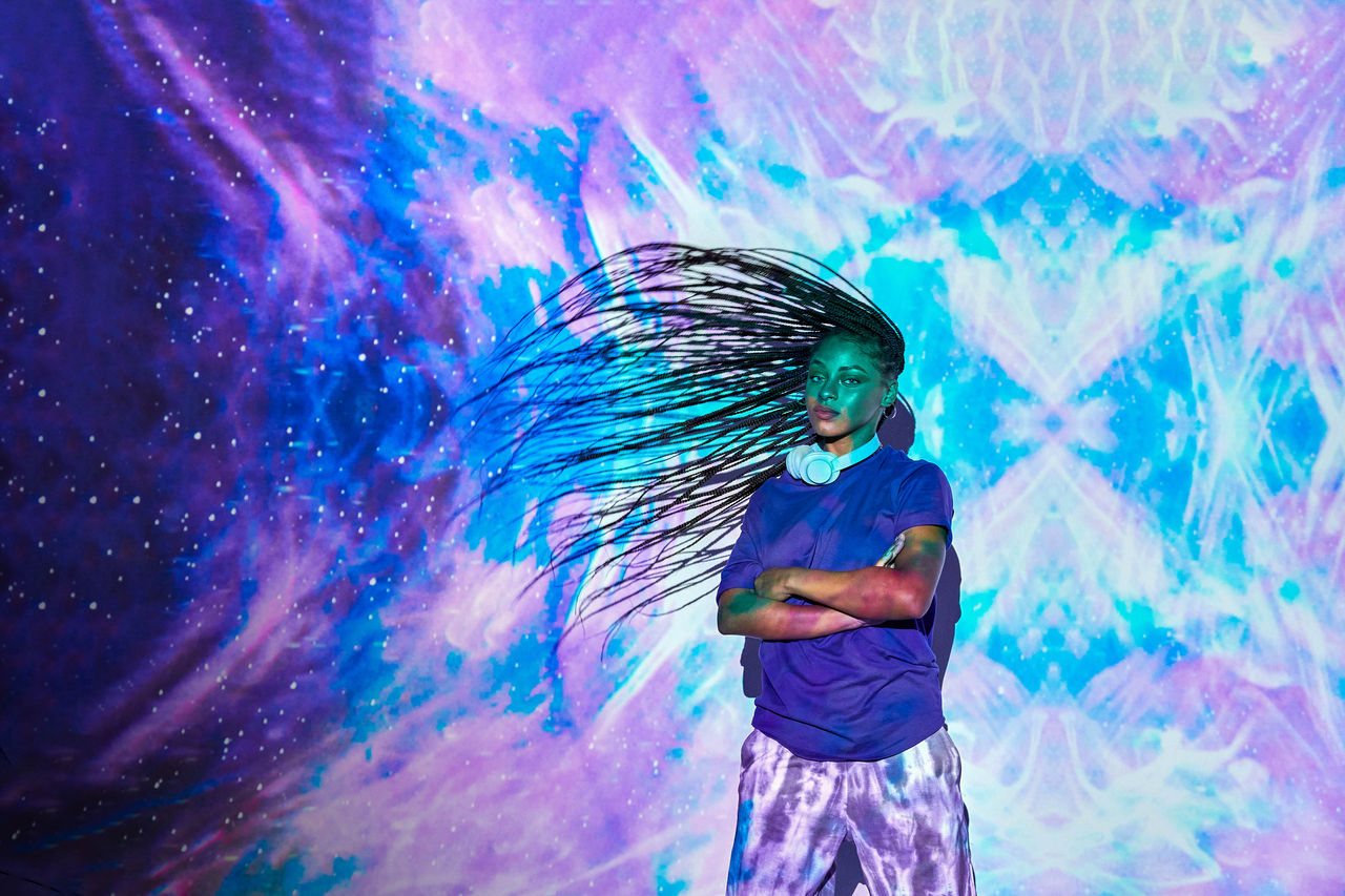 Lady with folded arms, tossing her braided hair, standing in front of a blue and purple projection